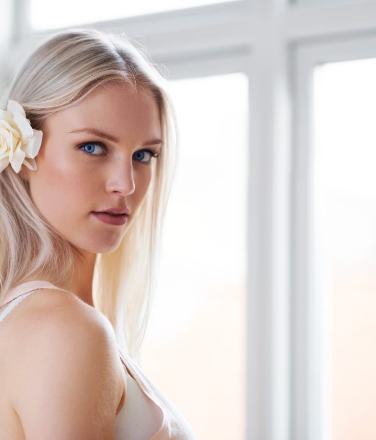 Portrait of an attractive woman with a white rose in her hair while standing in her bedroom.